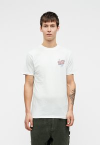 Mladý muž s krátkymi hnedými vlasmi, oblečený v bielom tričku s logom "Santa Cruz" a tmavozelenými nohavicami, stojí na jednoduchom bielom pozadí.