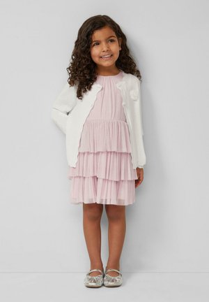 Fille souriante aux cheveux bouclés portant un cardigan blanc, une robe rose à volants et des chaussures argentées, debout devant un mur gris clair uni.