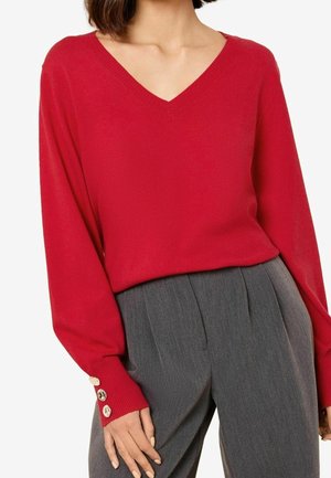 Frau trägt einen roten V-Ausschnitt-Pullover mit geknöpften Manschetten und eine hoch taillierte graue, plissierte Hose vor weißem Hintergrund.