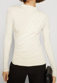 Krämfärgad långärmad topp med en asymmetrisk design, rynkad textur och hög krage, i kombination med svarta byxor och en strukturerad clutchn.