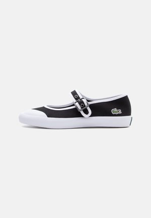 Black and white Mary Jane sneaker with buckle strap, white sole, and green Lacoste crocodile logo on the heel side.