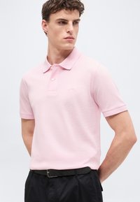 Νέος άνδρας που φοράει ροζ ανοιχτό πόλο πουκάμισο μέσα σε μαύρο παντελόνι με μαύρη ζώνη, κοιτάζοντας προς το πλάι με φόντο μια απλή επιφάνεια.