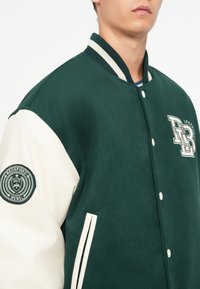 Grøn og creme varsity jakke med en blød tekstur, ribbet krave og lap på ærmet. Har hvide accentstriber og logo.