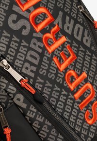 Czarny plecak z wyrazistym pomarańczowym haftem i szarym, powtarzającym się logo. Zawiera kieszeń zapinaną na zamek z pomarańczowymi uchwytami oraz teksturowaną powierzchnię.