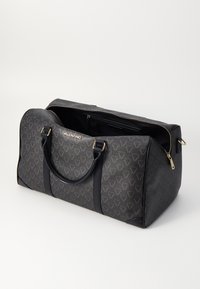 Μαύρη τσάντα duffle με υφή και χρυσές μεταλλικές λεπτομέρειες. Διαθέτει δύο δερμάτινες λαβές και ευρύχωρο, επενδεδυμένο εσωτερικό με κλείσιμο φερμουάρ.