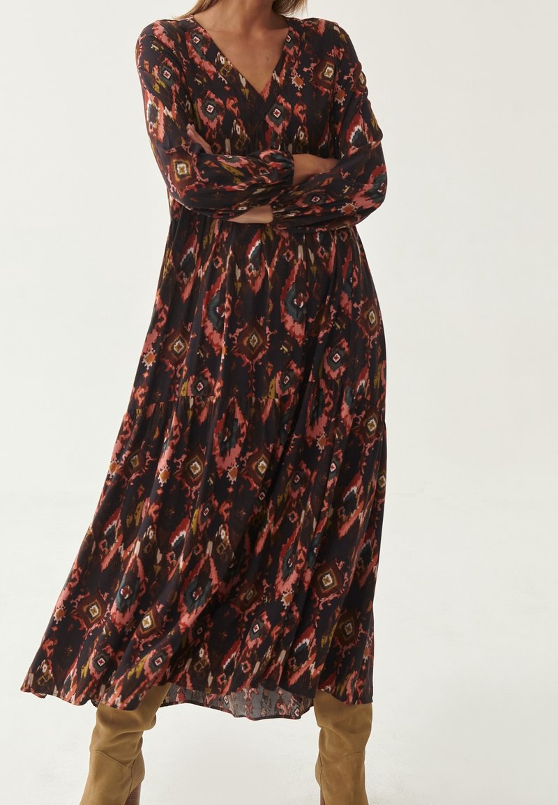 Femme portant une longue robe sombre à motifs avec des formes géométriques rouges et brunes, debout les bras croisés et des bottes hautes couleur beige.