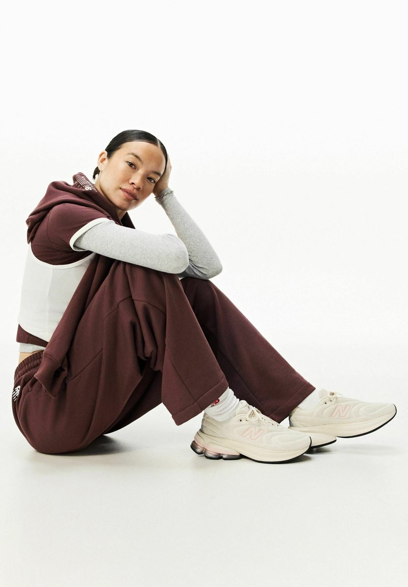 Donna con tuta bordeaux seduta a gambe incrociate sul pavimento, con la testa appoggiata sulla mano, indossa sneakers bianche e una maglia grigia a maniche lunghe sotto la felpa con cappuccio.
