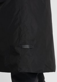 Abrigo negro hecho de tela suave y resistente al agua. Presenta una silueta estilizada con un detalle de correa abotonada cerca del dobladillo.