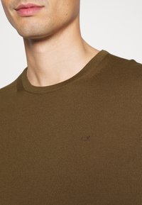 Khaki zelena pletena majica s okruglim izrezom i suptilnom teksturom. Ima mali izvezeni logo "CK" na prednjem dijelu.