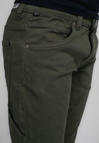 Olijfgroene cargo broek van een gestructureerde stof, met meerdere zakken, zichtbare stiksels en metalen hardware accenten.