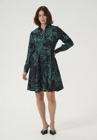 Das Hemdkleid besteht aus dunkelblauem Stoff mit grünem Blumenmuster, hat lange Ärmel, eine knöpfbare Vorderseite und einen ausgestellten Rock. Schwarze spitze Schuhe.