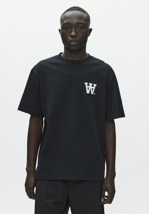 Ung mand med mørk hud iført en løs sort t-shirt med en hvid dobbelt "W"-logo på venstre bryst, stående mod en ensfarvet hvid baggrund.