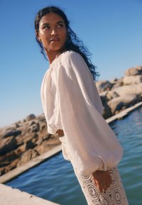 Light cream blouse with puff sleeves and a round neckline, paired with a white crochet skirt featuring circular patterns, set by water.