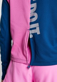 Langärmliges Sportoberteil mit pinken und blauen Farbblöcken, strukturiertem Stoff und einem großen grauen Logo an der Seite, mit einem Reißverschlussdetail.