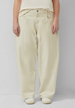 Person trägt locker sitzende beige Hosen mit Fronttaschen und weiße Sneakers, steht vor einem einfarbigen hellgrauen Hintergrund.