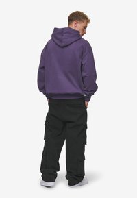 Sweatshirt purpuriu cu glugă, cu o croială lejeră, asociat cu pantaloni cargo negri, largi, cu buzunare, și adidași albi.