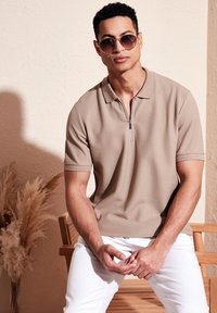 Bežs polo krekls ar rāvējslēdzēju, īsām piedurknēm, ar teksturētu audumu; kombinēts ar baltām biksēm un apaļām saulesbrillēm. Minimalistiska vide.