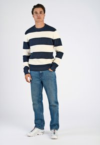 Blauwwitte gestreepte ribbelsweater met ronde hals, gecombineerd met blauwe spijkerbroek en witte sneakers, tegen een effen achtergrond.