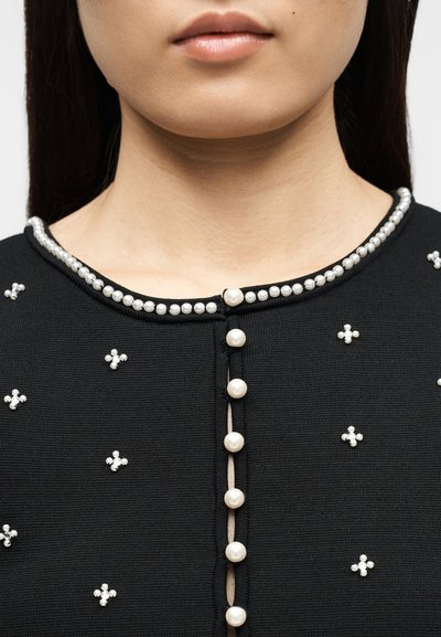 Γυναίκα που φορεί ένα μαύρο ένδυμα με κουμπιά από πέρλες κατά μήκος του κέντρου και μικρές ομάδες από πέρλες κεντημένες στο ύφασμα κοντά στη λαιμόκοψη.