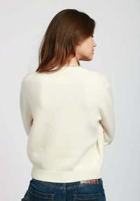 Donna con lunghi capelli castani, che indossa un maglione crema e jeans blu, in piedi con le braccia incrociate, di spalle rispetto alla fotocamera.