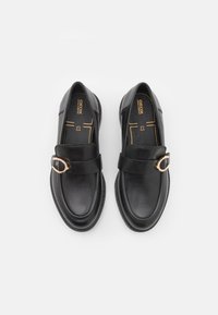 Mocasines de cuero negro con punta redonda y una hebilla decorativa en tono dorado. Textura suave y detalles de costura en la suela.