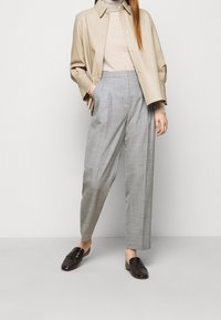 Veste en cuir beige sur un pull léger, associée à un pantalon à carreaux gris et des mocassins foncés, mettant en avant une coupe ample et un design ajusté.
