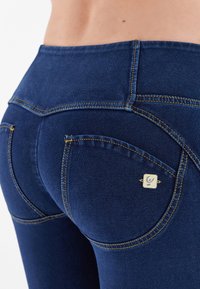 Jeans de mezclilla azul oscuro con costuras doradas, que presentan bolsillos traseros curvados y un pequeño acento metálico en la parte derecha cerca del bolsillo.