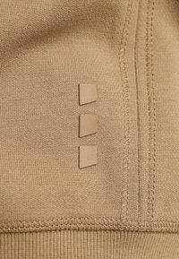 Gros plan d’un tissu beige avec trois petites pièces en caoutchouc en forme de trapèze alignées verticalement près d’une couture.