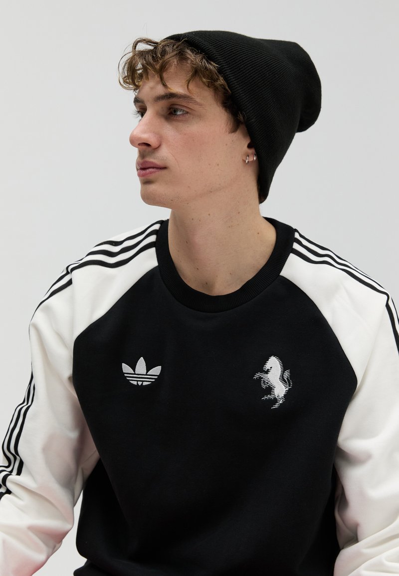 Schwarz-weißes Sweatshirt mit Rundhalsausschnitt, drei gestreiften Ärmeln, einem Adidas-Logo und einem aufgedruckten Pferdemotiv. Schwarze Beanie-Mütze.