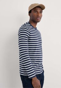 Camisa de rayas azul marino y blanca de manga larga con cuello redondo, combinada con jeans oscuros y una gorra beige. Diseño simple y casual.