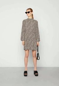 Mulher vestindo um vestido mini de manga longa com estampado de leopardo, sandálias pretas de plataforma, bolsa preta e óculos de sol estilo gato pretos, em pé contra uma parede branca.