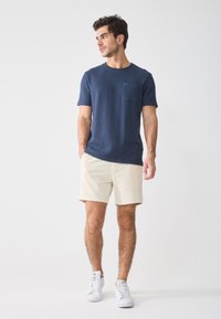 T-shirt navy a maniche corte con texture e taschino sul petto, abbinato a pantaloni corti beige e sneakers bianche, in piedi davanti a uno sfondo neutro.