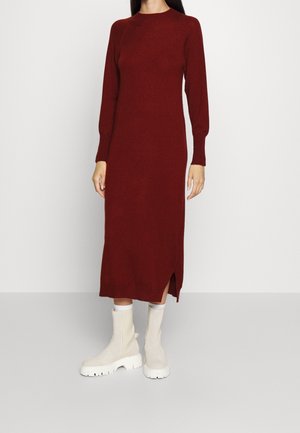 Kvinde iført en langærmet, ankellang strikket kjole i mørkerød med sideslidser, kombineret med hvide chunky ankelstøvler.