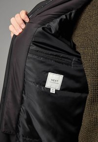 Schwarze Pufferjacke mit glatter Innenausstattung. Etikett sichtbar mit "NEXT TAILORING" und Größe M. Außenschicht ist in einem strukturierten Dunkelgrün.
