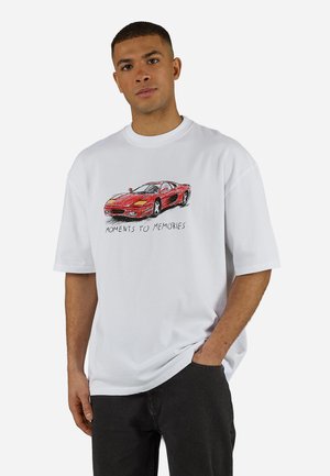 Nuori mies, jolla on valkoinen t-paita, jossa on punaisen urheiluauton luonnos ja teksti "MOMENTS TO MEMORIES", seisoo käsi taskussa.