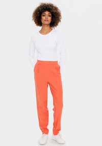 Orange Hosen mit Seitentaschen, kombiniert mit einem körperbetonten weißen Langarmshirt. Weiße Sneakers vervollständigen das Outfit. Sanfte Textur, maßgeschneiderte Passform.