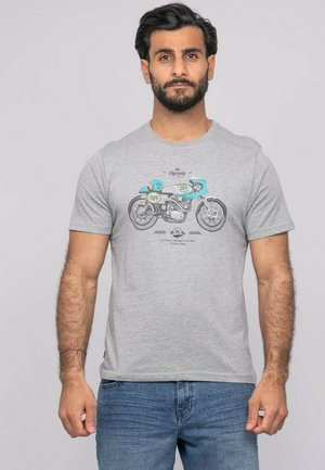 Homme aux cheveux foncés et à la barbe, portant un t-shirt gris avec un motif de moto vintage bleu et noir, debout devant un fond uni.