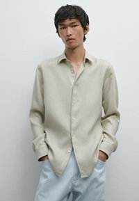 Light beige long-sleeve shirt in a woven fabric, featuring a button-down front, relaxed fit, and pointed collar, paired with light blue pants.