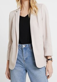 Blazer rose clair avec une coupe ajustée, col à revers cranté et manches longues, associé à un t-shirt noir et un jean bleu taille haute.