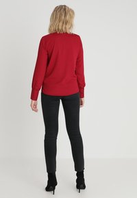 Rød langærmet skjorte med knapdetaljer ved manchetterne, parret med sorte slim-fit jeans. Stoffet virker glat og let.