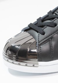 Black leather sneaker featuring a metallic silver toe cap with ridges, textured white sole, and black laces. Smooth and reflective surfaces.