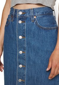 Niebieska jeansowa spódnica midi z zapięciem na guziki z przodu, białe metalowe guziki i kontrastowe przeszycia. Posiada dwie przednie kieszenie i dopasowaną talię.
