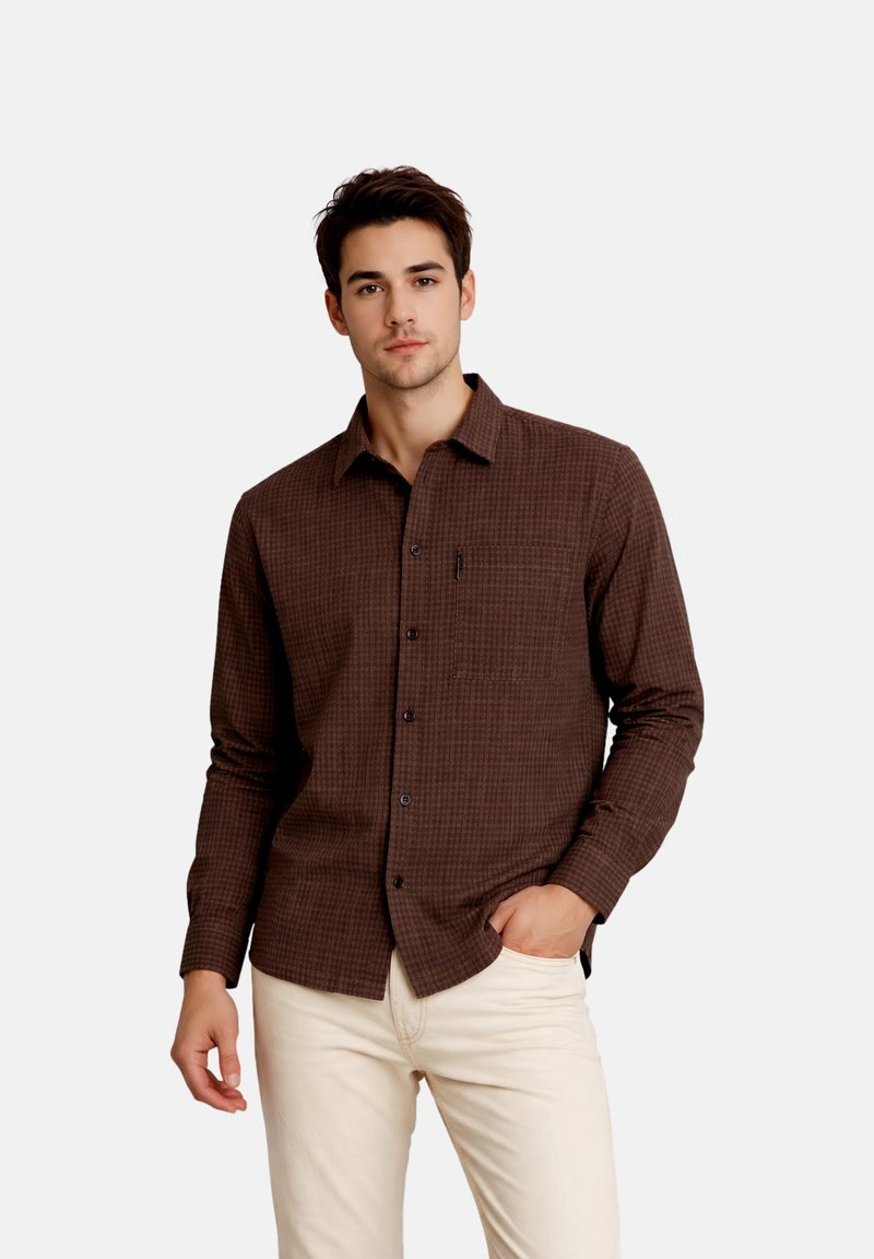 Jeune homme portant une chemise à carreaux marron à manches longues et un pantalon beige clair, debout avec une main dans la poche devant un fond uni.