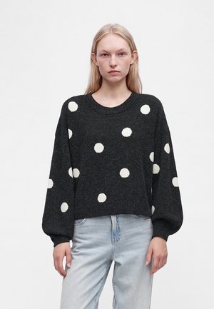 Jeune femme aux longs cheveux blonds porte un pull noir à gros pois blancs et un jean bleu clair, debout devant un fond uni.