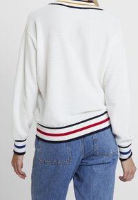 Vit stickad tröja med ribbade ärmslut och nederdel. Randiga detaljer i marinblått, rött och blått. Kombinerad med högmidjade denimjeans.