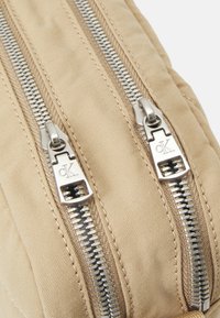 Beżowa torba materiałowa z dwoma srebrnymi zamkami błyskawicznymi z wyspecjalizowanymi uchwytami. Teksturowana powierzchnia z widocznymi szwami. Minimalistyczny design.