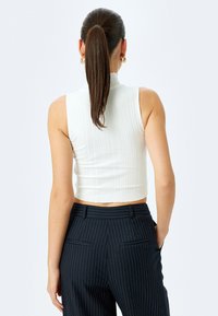 Cropped, ribbed white sleeveless top with a high neckline. Paired with navy pinstriped trousers featuring back pockets.