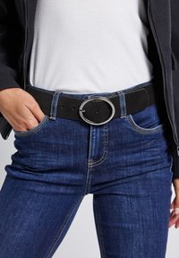 Osoba v modrých džínách s černým pásem, na kterém je velká kulatá stříbrná přezka, ruka v kapse, bílá košile a tmavá bunda.