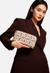 Portafoglio beige con motivo geometrico marrone, parti in metallo dorato e chiusura con cerniera, tenuto in una mano contro un blazer marrone scuro.