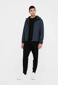 Giacca blu navy con cerniera e cappuccio, dotata di due tasche frontali e una texture liscia, abbinata a una felpa nera e pantaloni neri.